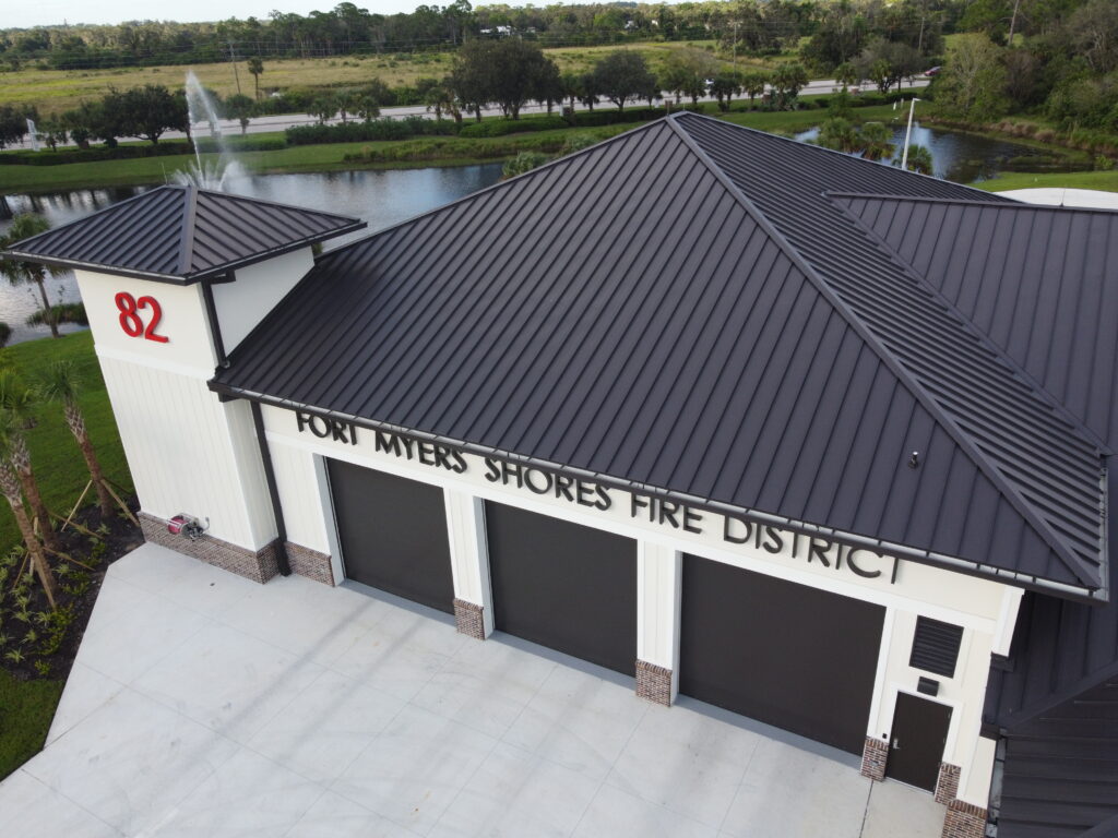 Fort Myers Shores Fire District building featuring a modern metal roof, large garage doors, and prominent address number 82, surrounded by landscaped greenery and water features.